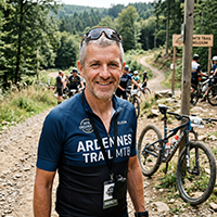 Realistic portrait of a 52-year-old Belgian man, mountain bike organizer, outdoors on a dirt path with a mountain bike visible in background, short grey hair, friendly smile, wearing a bike jersey and sunglasses, bright daylight, medium shot, natural and approachable expression.
