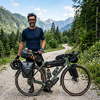 Realistic portrait of a 41-year-old Slovenian man, gravel bikepacker, standing next to a loaded gravel bike on a forest road, stubble, focused but friendly expression, cycling glasses, helmet in hand, green alpine background, outdoor natural light.