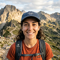 Realistic headshot of a 28-year-old Spanish woman hiker, outdoor in the Pyrenees mountains, wearing a hiking backpack and a cap, smiling warmly, sunlit rocky landscape behind her, natural colors, joyful expression, medium close-up.